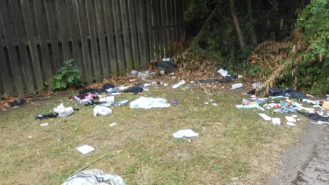 Rubbish is strewn around a grassy piece of land close to a wooden fence. The litter can be sen in several areas.
