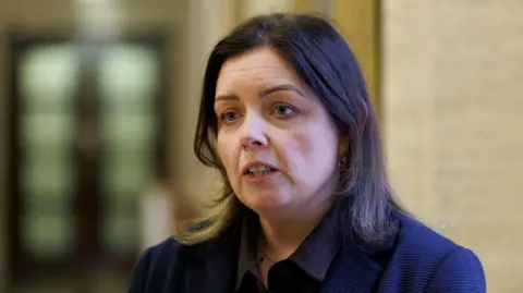 PA Media Deirdre Hargey has shoulder-length dark hair. She is wearing a dark, blue-ish jacket and shift.