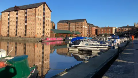 Several docked boats, with large red-brick warehouses behind them. The sky is bright blue, it's a sunny day. 