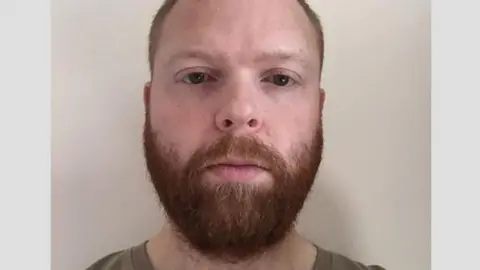 A police mugshot of Paul Chowles wearing a brown T-shirt and standing against a white background. He has a full beard and short red-brown hair, and is looking at the camera with a blank expression. 