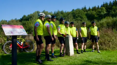 BBC Eight cyclists in lime green gilets and cycling gear, standing in the sunshine at kilometre zero in Switzerland 