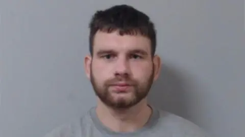 PSNI Marc Doak, pictured in custody. He has dark hair and a beard and is wearing a grey top.