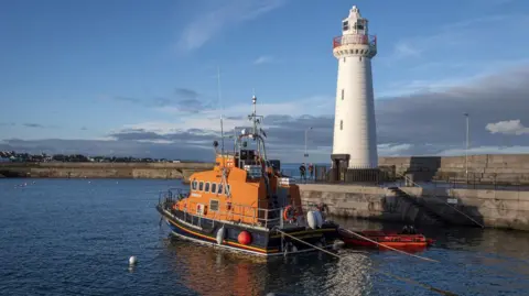 Rossographer/Geograph Donaghadee Harbour with a white lighthouse at the end of one half of the harbour walls. In front is an orange and navy blue lifeboat. The harbour waters are calm and dark blue. Beyond the harbour walls is the sea and an expanse of low stratus clouds. It's feels like a sunny evening.