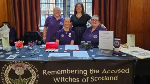 RAWS A group of four women sit behind a table - three are wearing purple tops and one is in a black dress. On the table are various leaflets and booklets. A banner draped over the table reads REMEMBERING THE ACCUSED WITCHES OF SCOTLAND.