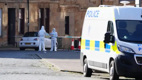 PA Media Forensic officers in white suits stand outside a tenement block, next to police tape. A white police van is in the foreground on the right of the picture.