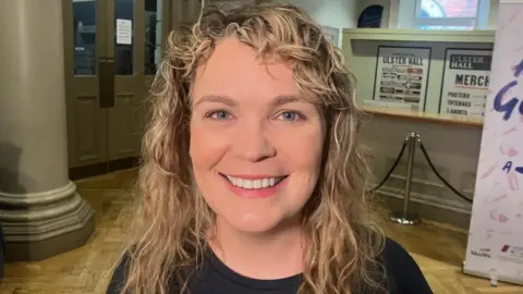 Has curly blonde hair and is smiling at the camera. She is wearing a black top.