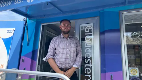 Richard Barrow, in a checked shirt, stands in front of the door into the NHS Gloucestershire mobile medical unit