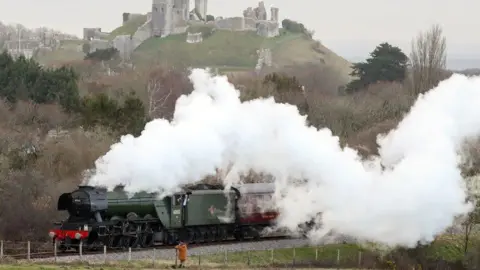 The dark green Flying Scotsman steam locomotive emerges from a cloud of white steam, pulling a line of maroon-coloured carriages, as it passes below the hilltop ruins of Corfe Castle.