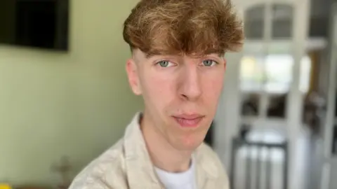 Nikki Fox/BBC Third year student midwife Jack Foreman looks at the camera. You can see his head and shoulders. He has a narrow face with blue eyes. He is wearing a cream jacket and white t-shirt. His hair is wavy and mousey and his hair is shaved underneath. He is standing in his kitchen.