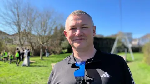 A man with short cropped hair is smiling at the camera. He is standing in a park with trees and houses in the background