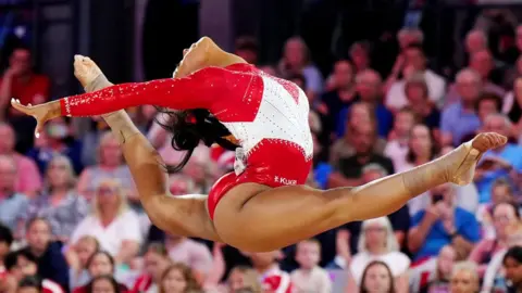 PA Media Image of a gymnast wearing a red and white leotard. She is pictured in mid-air in a scissor leap.