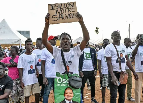 AFP/Getty Images Apoiadores de Jean-Louis Billon em camisetas brancas seguram um cartaz em apoio a ele que diz “Bilon é nosso salvador” em francês.