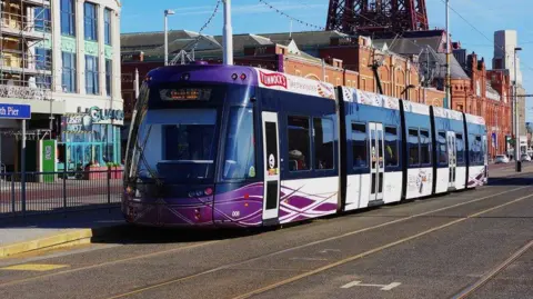 A Blackpool tram, with purple and grey livery, travels along the promenade in front of arcades and the Blackpool Tower