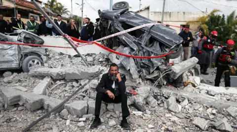 Man sitting beneath wrecked car looking at the camera with wrecked car behind him