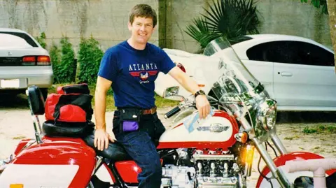 Gordon Castle Gordon is sitting on a huge red Honda Valkyrie with a big windscreen. He is wearing a blue t-shirt with the word Atlanta on it and blue jeans.