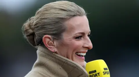 Getty Images A blonde-haired woman with a happy face, wearing a beige jacket with the collar turned up. She is smiling and looking off into the distance while holding a microphone. 