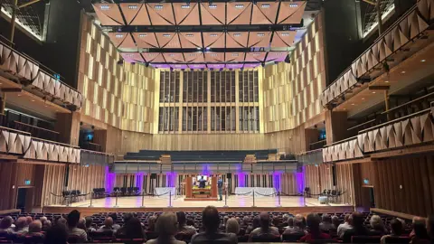 Image of a concert venue, with a seated audience in the foreground watching a man on stage playing a vast pipe organ
