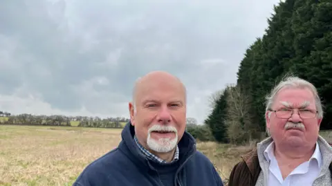 Claire Cavanagh Two men, campaigners Martin Thomas and Steve Reade, stand in front of a field. Just their heads and shoulders can be seen. The field is lined with trees. 