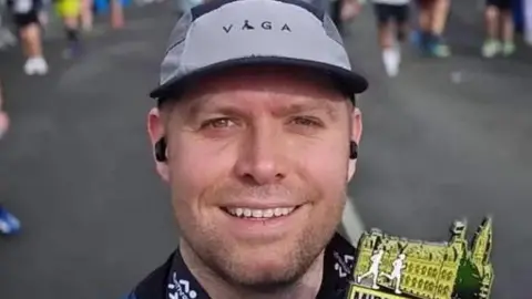 Instagram/Carmel College PC Blades takes a selfie with a medal for completing a run in Middlesbrough. He has neat stubble and is smiling, with a grey running cap on his head.