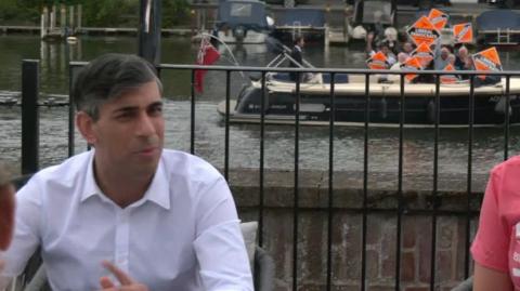 Rishi Sunak speaking while a boat of Liberal Democrats sail past with signs