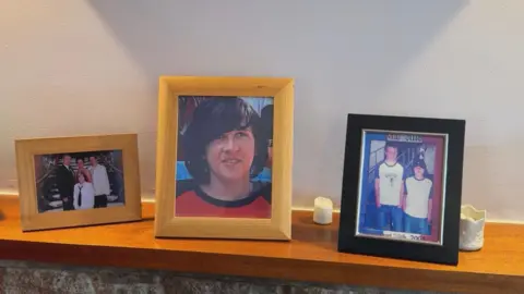A mantelpiece with framed photographs of a young boy, possibly a teenager. A couple of candles are on the mantelpiece.
