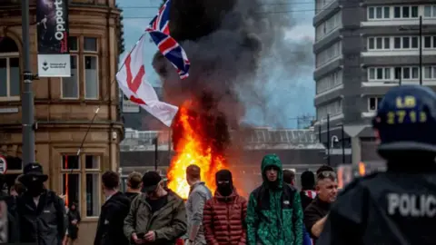 Getty Images Crowds gather in front of a burning building, with riot police visible in the corner of the image