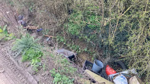 Laura Coffey/BBC A ditch showing a tyre, plastic, gas canister, plastic bags and shrubs and hedges.