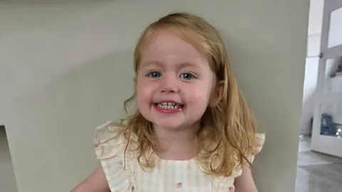 McGeoch Family A young girl with blond hair smiles into the camera while wearing a bright white and light pink dress. She is leaning against a wall.