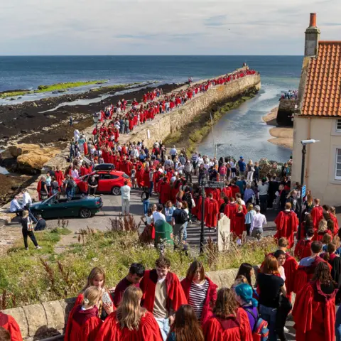 University of St Andrews  Lots of people in red gowns walking across the long pier