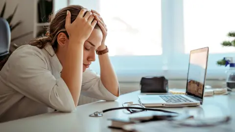 A woman sits at a desk facing her laptop with her head in her hands