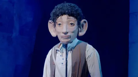 A puppet of a giant with black hair, big ears, grey shirt and brown waistcoat