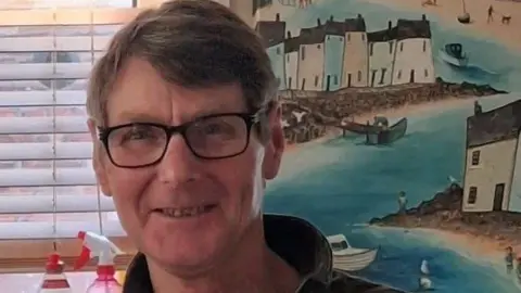 Family Handout A man with brown hair, dark rimmed glasses and a dark jumper smiles into the camera. He is positioned in front of a window with a colourful picture depicting a seaside harbour visible over his shoulder.