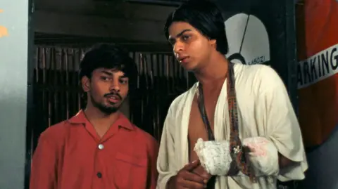 The Film Heritage Foundation A young Shah Rukh Khan (on the right), seen in a white robe and a plaster and a sling in his right hand which seems to be broken. He is standing next to a shorter man, wearing a red t-shirt.