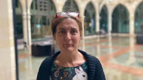 Laura Coffey/BBC Tina Matthew with short brown hair and brown glasses on her head, wearing a blue cardigan and a blue dress with flowers and a rabbit on it. She is standing outside Northampton Guildhall, the stone archways of which are visible behind her