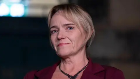 PA Media A portrait of a woman, shown from the shoulders up, with a dark background and soft lighting. She has medium-length hair that is neatly styled.