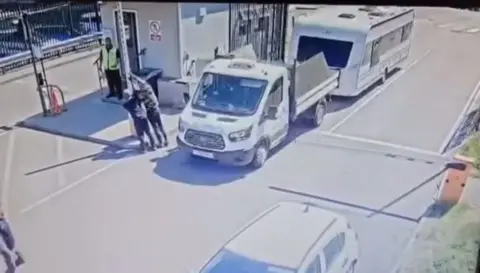 onlyurbexbh/TikTok A white tipper truck towing a caravan passes a gatehouse as two men hold up the barrier. A security guard in a yellow jacket stands on the stop of his gatehouse.