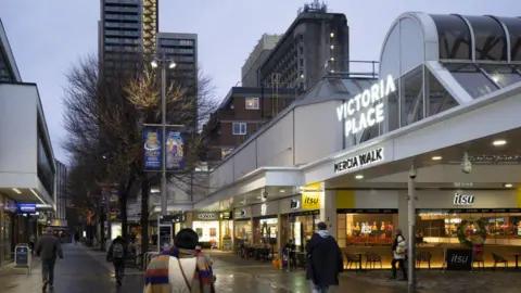 Commercial Way in Woking on a dark, overcast day. High street brands including Itsu, Howden and Betfred have signs outside their shopfronts. A large, white shopping centre with a sign that reads 'Victoria Place' is seen on the right. There are tow high-rise buildings in the background.