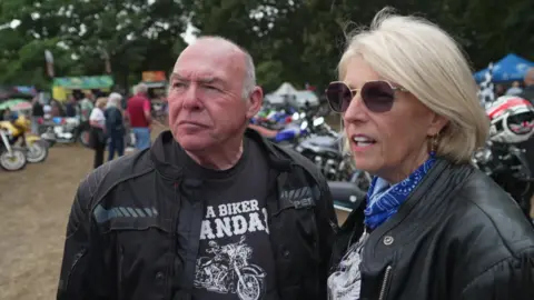 A man and a woman stood in the middle of a grass field, with motorcycles behind them, looking to the left of the camera. The man is bald and is wearing a black T-shirt with white branding on and a black jacket. The woman has a short, blonde bob and is wearing a black jacket and a blue and white scarf. 