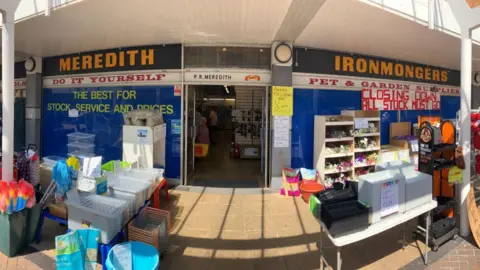 Image of the shopfront of an ironmongers with merchandise including dusters, mops, plastic storage crates and seed trays stacked outside