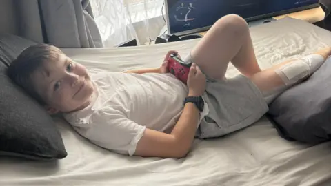 Nine-year-old Theo Rees lying in bed at home holding a red Xbox remote. He is wearing a white t-shirt and grey jogging shorts and has a big plaster on his right leg.