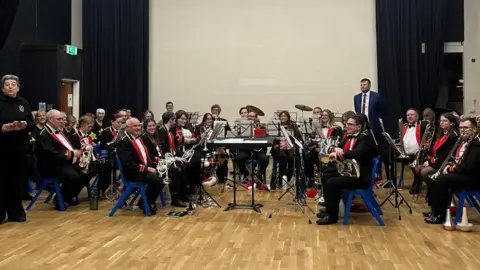 Image shows band members at a rehearsal in a hall. The band players are sat in a large semi circle and are wearing black jackets with a red opening and white shirts. They are also wearing black trousers and sat on blue plastic chairs. They are holding a range of brass instruments. The chairs are on a wooden floor and there is a white backdrop behind them. 