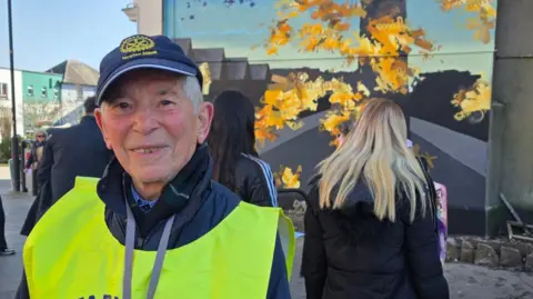 Ron Atkinson stood on the left wearing a dark blue jacket with a yellow hi-vis over the top. He is smiling at the camera and is stood in front of the large mural. Behind him is other people stood in front of the artwork. 