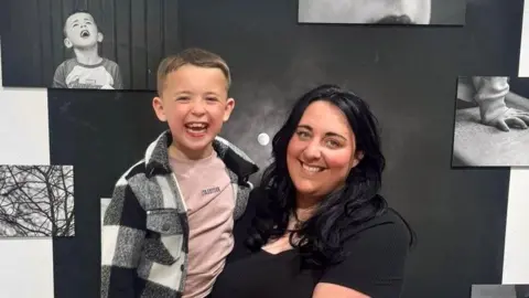 Bo Willow Harris, in a black top and leopard print skirt is holding her son Hendrix. They are both smiling at the camera while standing in front of black and white photographs attached on a white wall as part of the exhibition. Hendrix has short dark blonde hair and is wearing a black and white jacket, pink t-shirt, and a black trousers. His mother has long wavy black hair.