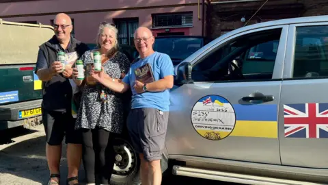 Phil Billingham Three people - two men and a woman - stand holding cans of beer and smiling at the camera. A car with Ukrainian flag livery and a Union Jack on its doors is to one side of them.