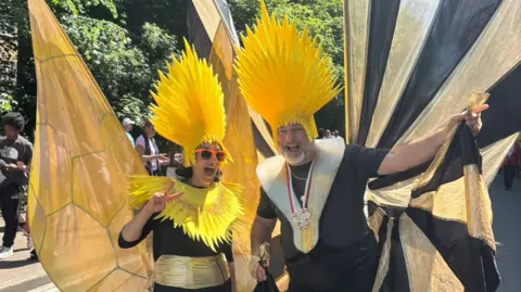 Macie Lewis/BBC A man and woman wearing black and gold carnival outfits complete with large wings pose for the camera. They are also wearing large yellow headdresses that rise up above their heads.