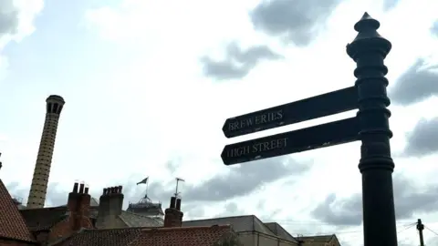 A signpost with two signs reading: Breweries and High Street. In the background, a view of rooftops and a light coloured brick chimney can be seen.