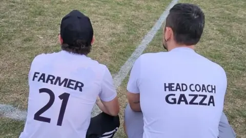 Handout Two men are seen from the back. They are crouching down on the touchline of a football pitch looking further into the centre of the pitch at what is going on. They are both wearing white T-shirts with dark writing on the back. One carries the name Farmer and also has the number 21 on it. The other T-shirt says Head Coach Gazza on the back of it.
