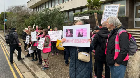 JULIETTE PARKIN/BBC About a dozen people, some wearing purple hi vis waistcoats and some holding up home made placards, stand outside Hove Trial Centre, a grey modern brutalist building.