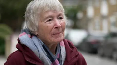 Sister Christine Frost is standing in a residential street, wearing a dark red coat and a grey and pink scarf 