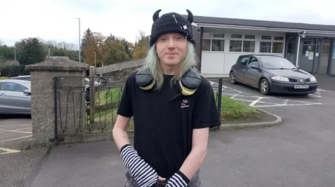 Man standing in a car park wearing headphones, a beanie with horns, a black t-shirt and white and black striped gloves 